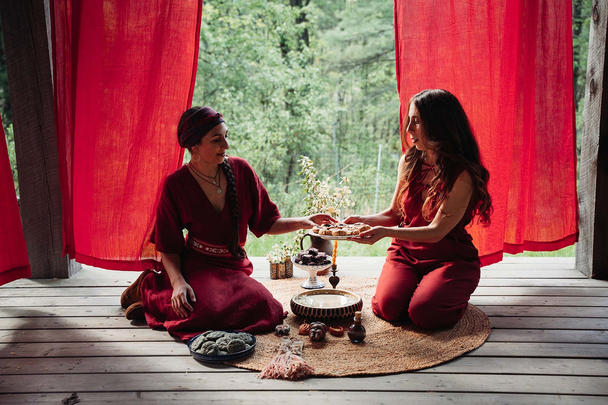 Two women in red outfits sitting on a wooden deck with red curtains and greenery in the background.