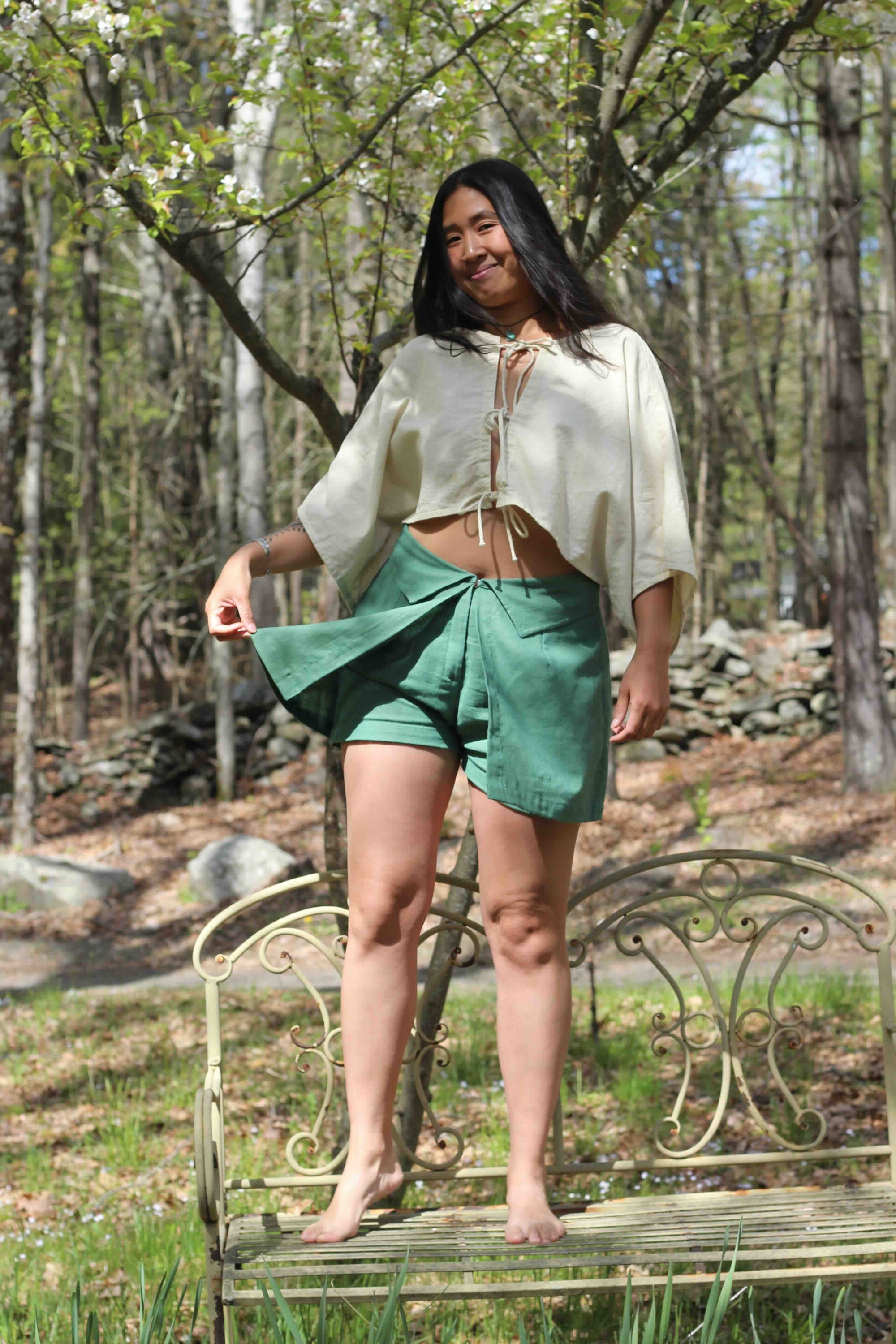 Woman standing on a bench outdoors wearing a white blouse and green shorts.