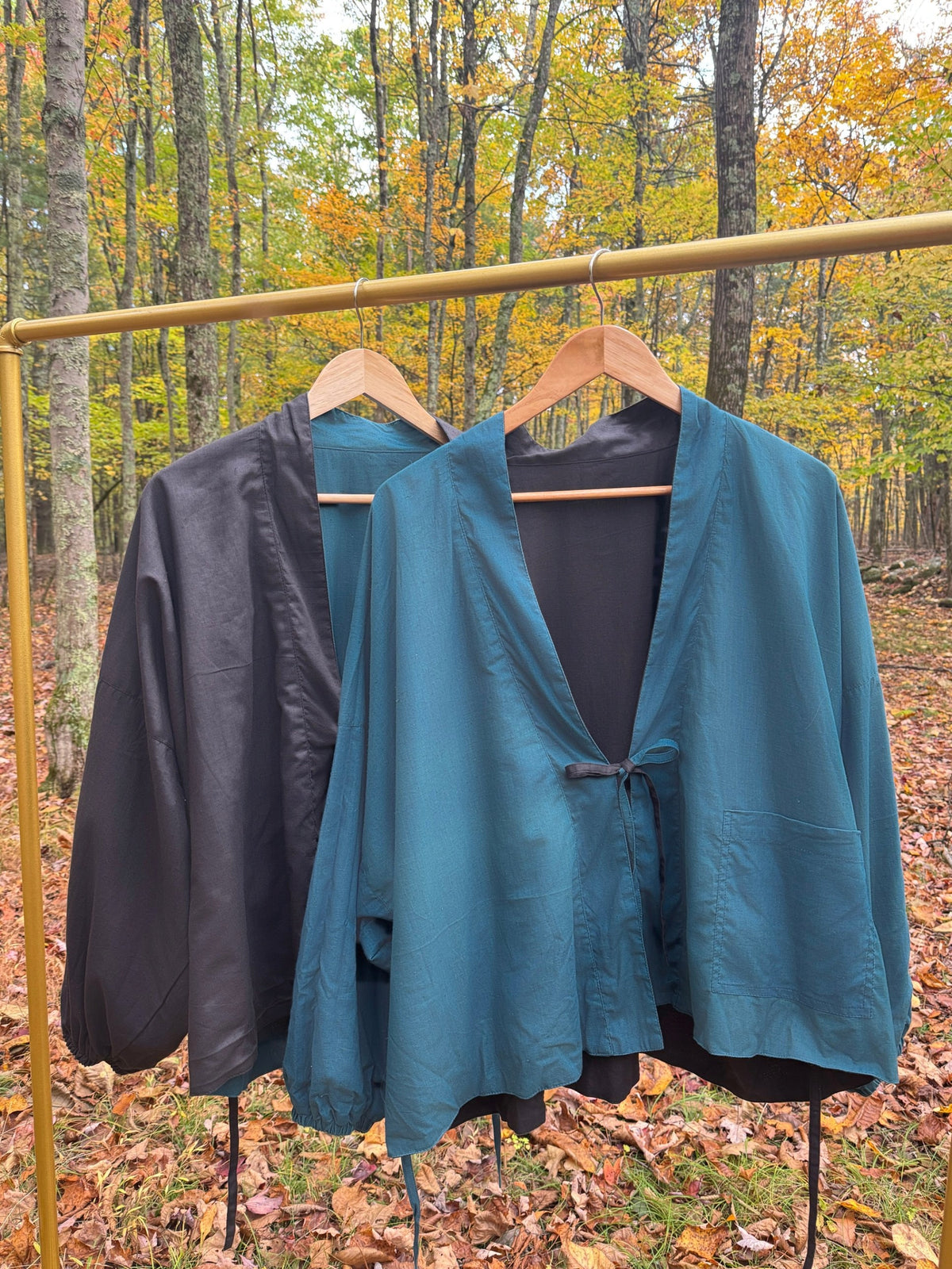 Black and teal sides of the reversible wrap jacket hanging on a gold rack in an autumn forest
